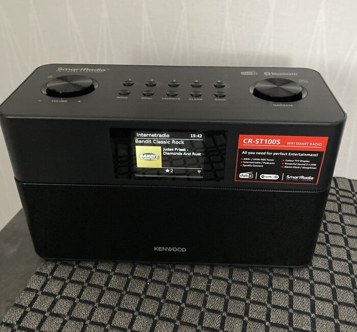 Kenwood CR-ST100S WiFi Smart Radio på ett bord, skärm visar 'Bandit Classic Rock', röd dekal synlig.