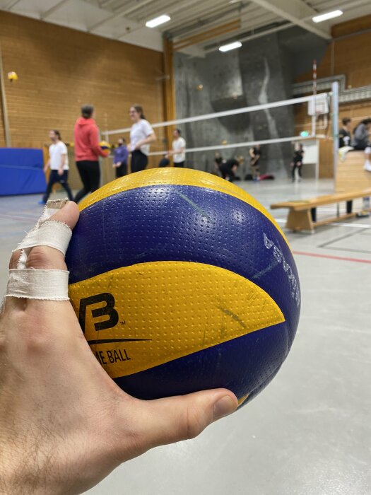 En närbild av en hand som håller i en blå och gul volleyboll i en sporthall med oskarpa spelare i bakgrunden.