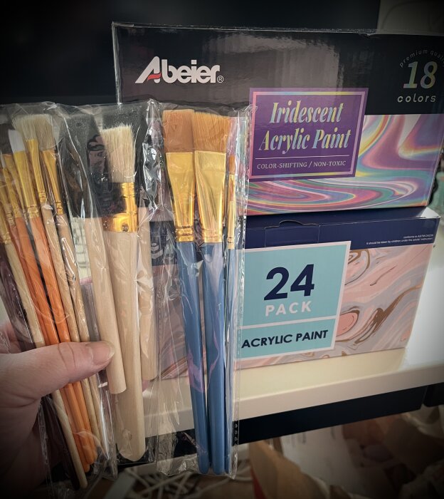 Hand håller paket med penslar och akrylfärger i förpackningar märkta "Iridescent Acrylic Paint" och "Acrylic Paint".