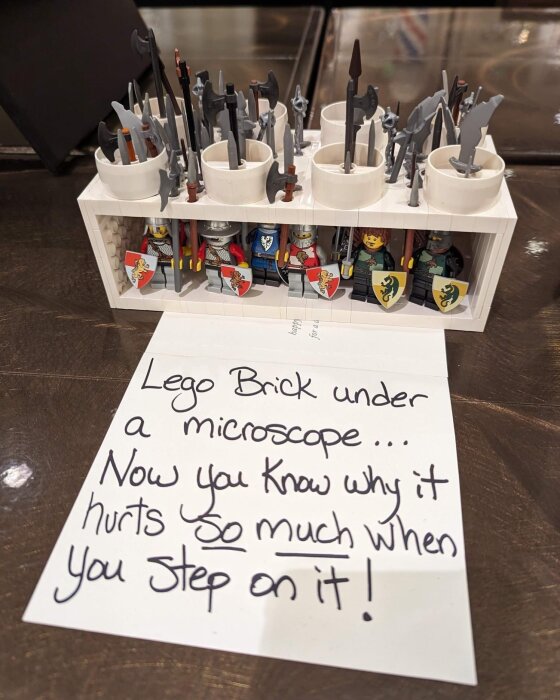 Legofigurer och vapen förvarade i en vit hylla med en humoristisk text på en lapp om Legobitar och smärta vid påtrampning.