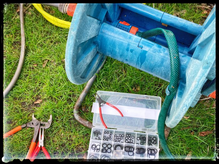 Öppen verktygslåda med olika storlekar packningar och en sidavbitare, intill blå plastsäckställning på gräs.