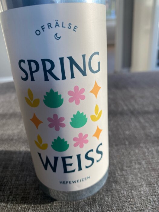 Flaska med etikett "SPRING WEISS Hefeweizen" i solljus, med färgglada blom- och lövmönster.