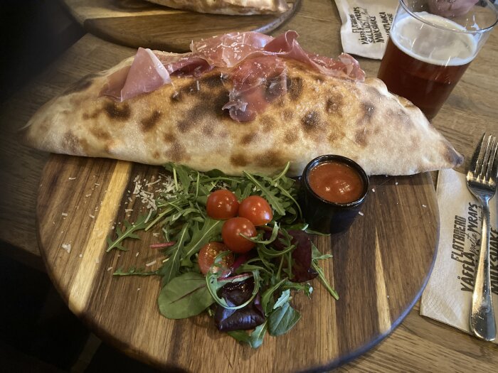 En välgräddad calzonepizza med lufttorkad skinka serveras på träbräda med sallad, körsbärstomater och tomatsås.