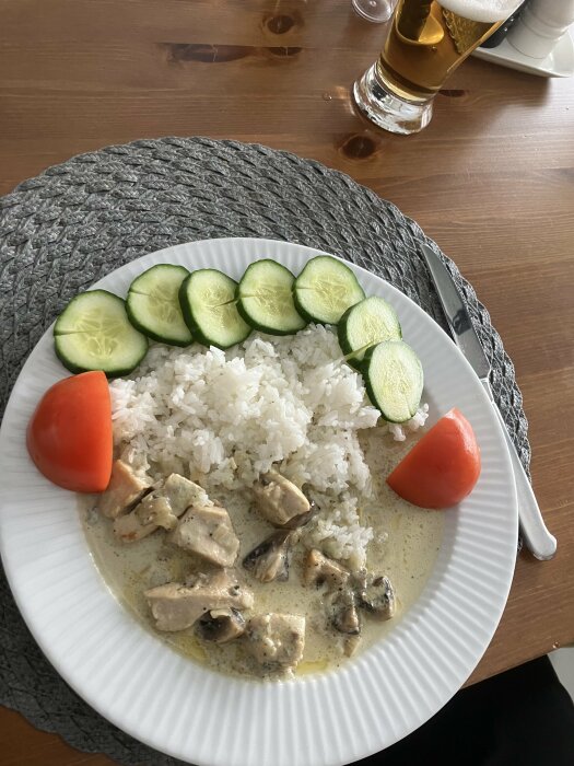 Tallrik med kycklinggryta gjord med ädelost och svamp, serverad med ris och gurkskivor, och en glas öl i bakgrunden.