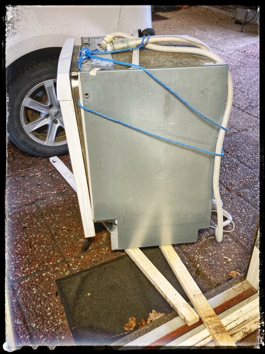 En gammal diskmaskin säkrad med blåa remmar på en pall nära en bil, redo för bortforsling.