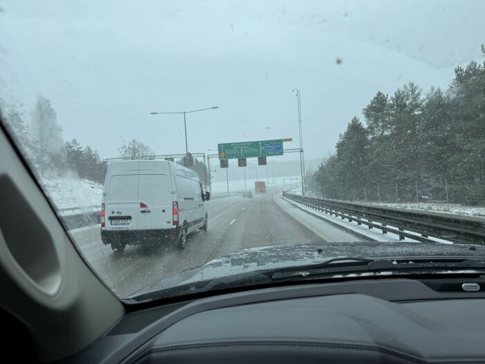 Vit skåpbil kör i vänsterfil på snöig motorväg under snöfall.