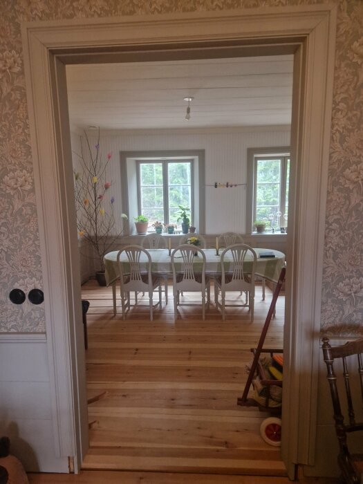 Vy genom dörröppning till matsal med trägolv, vitmålade möbler och blommigt tapetserade väggar.