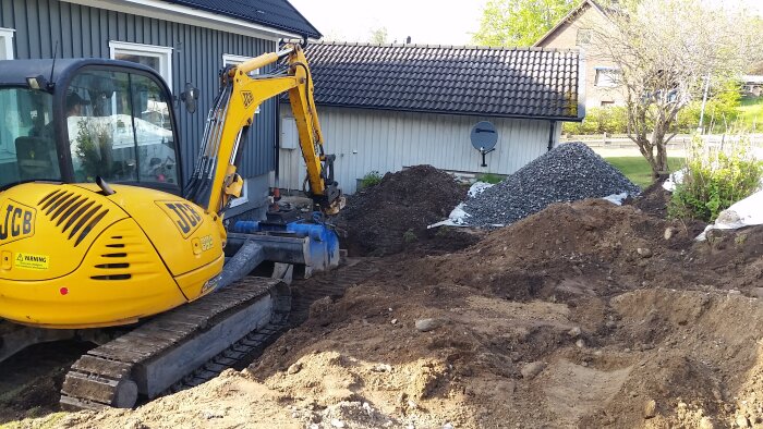 Grävmaskin vid grävarbete nära hus, brun sandig mark med sten och högar av grus och jord.