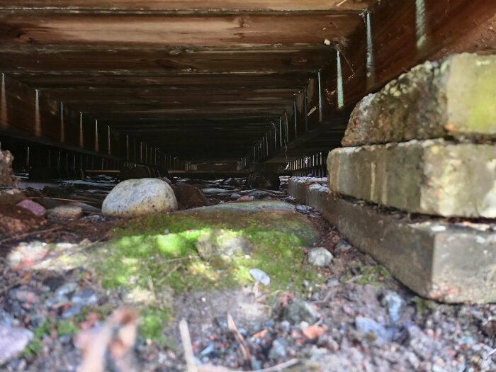 Vy under en byggstruktur där träbjälkar vilar på en blandning av stenplattor och stockar, ej nedgrävda.