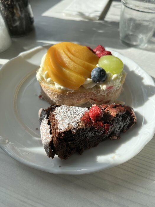 Desserttallrik med en bit kladdkaka toppad med bär och en bakelse med frukt och grädde.