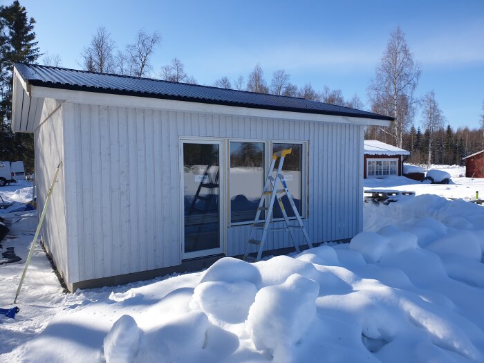 Attefallshus under konstruktion med fönster på långsidan, omgiven av snö och en stege utanför.