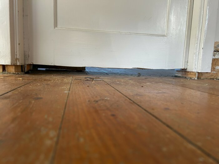 En sågad nedre del av en vit dörr ovanför ett gammalt trägolv som visar en glipa där golvet tidigare täckts av ett annat lager.