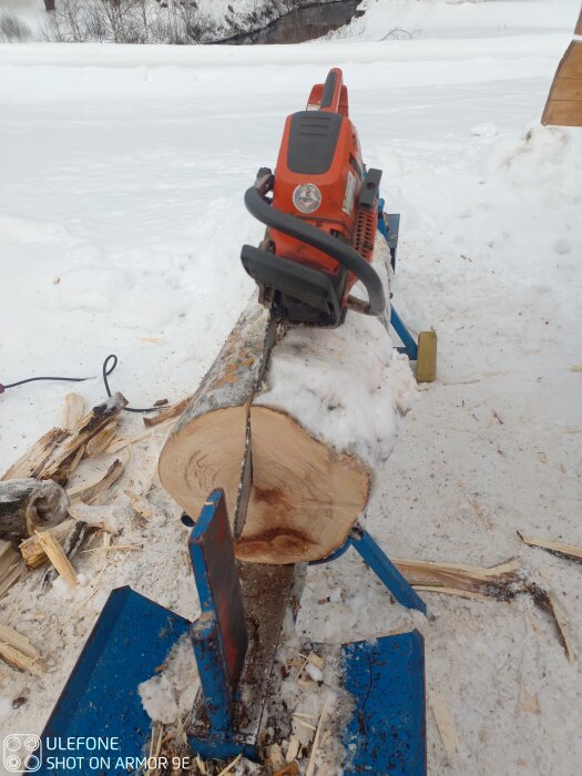 En motorsåg sågar igenom en trädstam som är placerad på en vedklyv utomhus i snön.