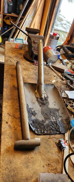 Ett brutet skyffelskaft bredvid bladet på ett smutsigt arbetsbord med verktyg och byggmaterial i bakgrunden.