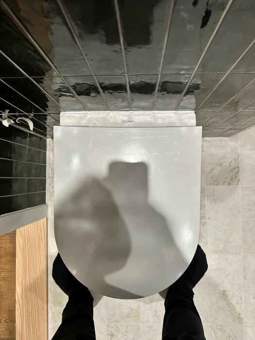 Uppifrån-bild på en snedställd toalettstol med ojämnt satta, spegelblanka kakelplattor i bakgrunden.