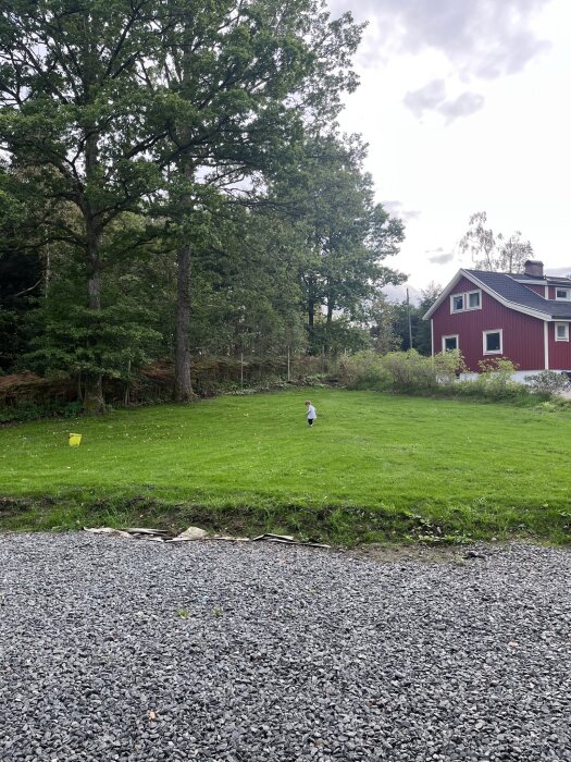 Spetsigt hörn av en tomt med grön gräsmatta, grus och träd i bakgrunden, med hus delvis synligt.