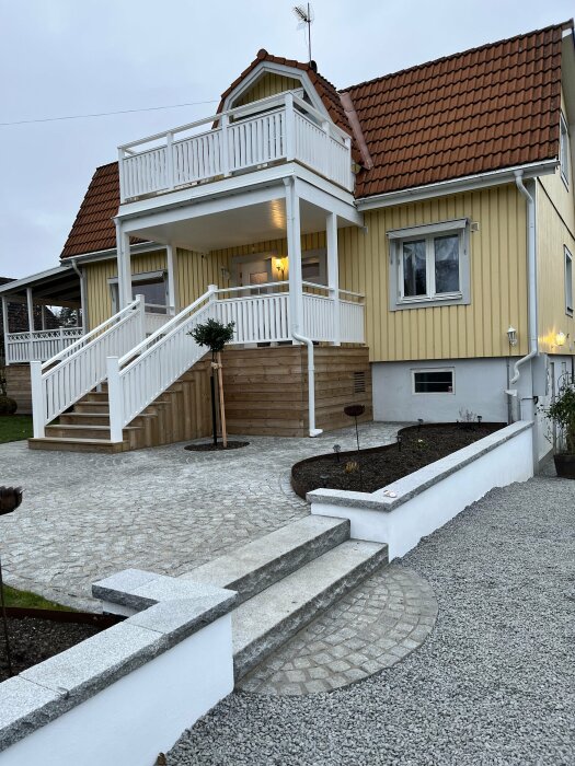 Ett gult hus med vit veranda och trapp som leder till en trädgård med vit mur, cortenkant och smågatsten.