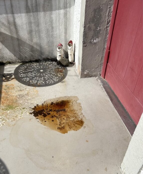 En entré med en röd dörr och en försänkning i betongen där vatten har samlats och lämnat en mörk fläck.