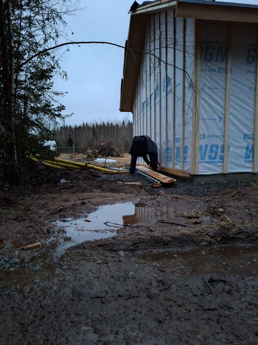 En person monterar vindpapp på ett halvbyggt hus med vindpapp och rullar vid fötterna på en lerig mark.