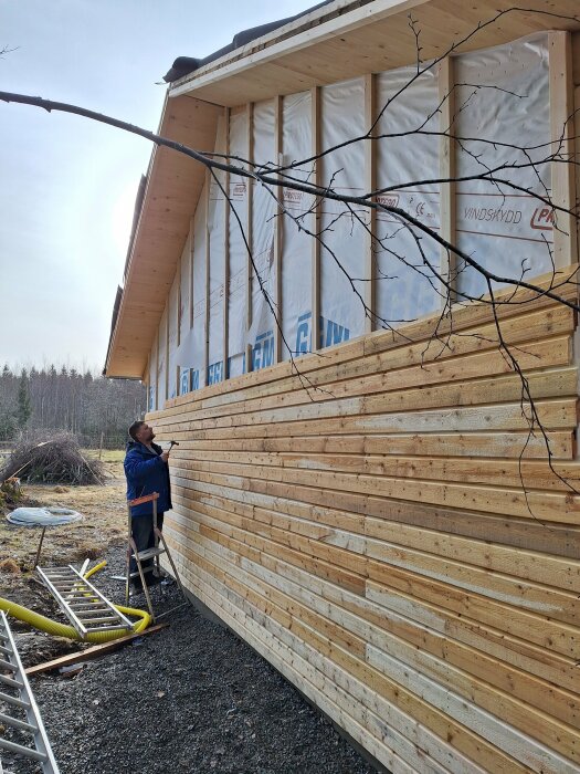 En person monterar en liggande träpanel på ett hus med vit vindspapp i bakgrunden och en stege på marken.