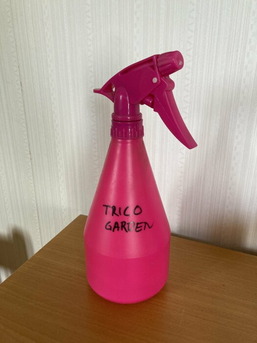 Rosa sprayflaska med texten "TRICO GARDEN" mot en tapetserad vägg.