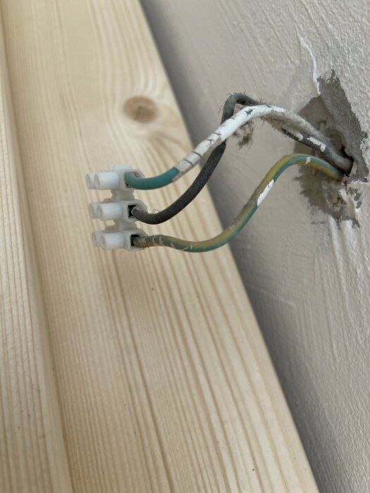 Lösa elektriska kablar med skarvhylsor sticker ut från ett hål i en vit vägg nära träreglar.