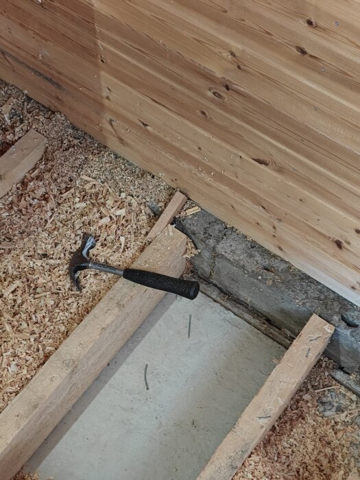 Betongplatta med uppreglat trägolv och sågspån, trävägg och hammare i renoveringsprojekt.