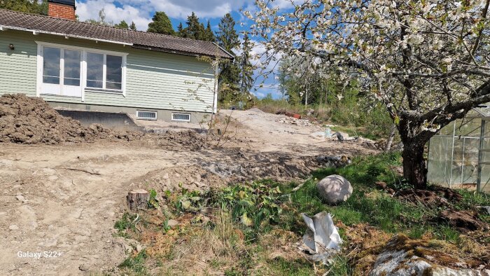 Källarrenovering på hus med synlig isodrän och utgrävd mark, omgivet av grönska och ett blommande träd.