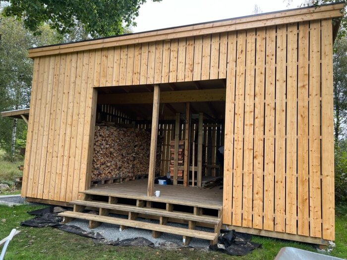 Träbod med öppen sektion för vedlagring, trappsteg och tak för virkesförvaring.