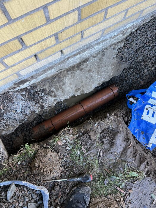 Uppgrävd dränering med synliga skador på ett rött tegelrör vid husgrund, omgiven av grus och jord.