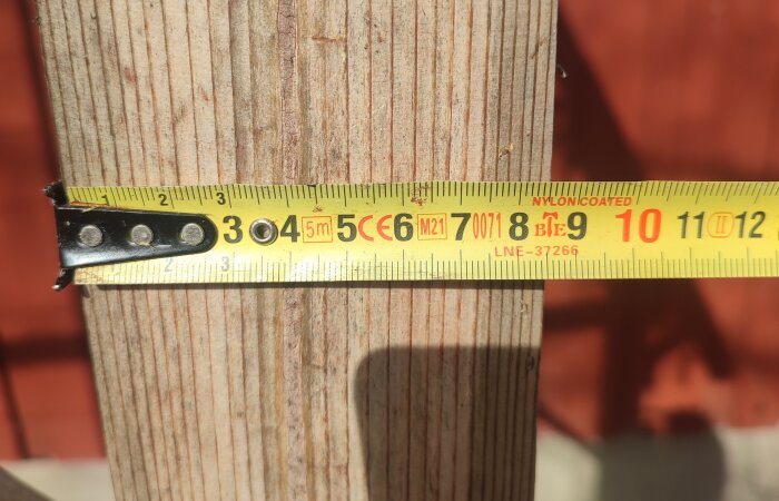 Måttband längs en träplanka som visar en mätning, delvis i skugga på en solig dag.