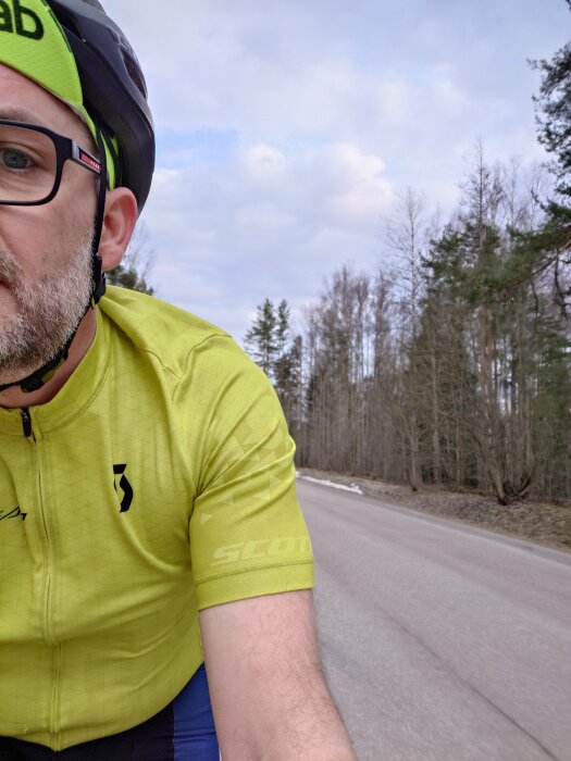 Person i gul cykeltröja och hjälm cyklar på en asfalterad landsväg med skog i bakgrunden.