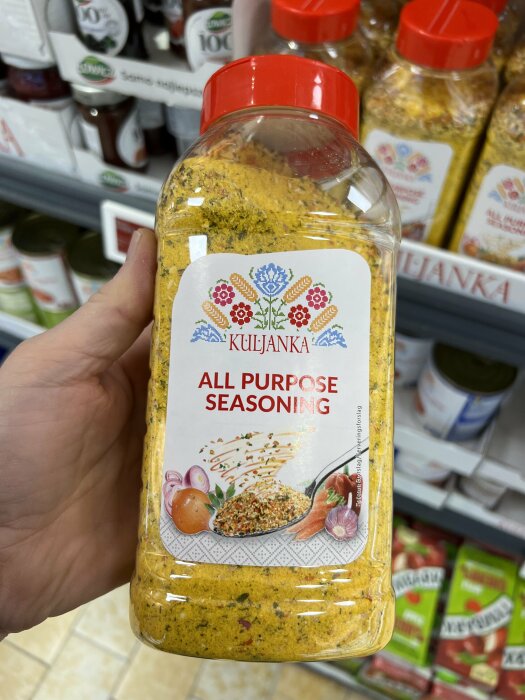 Hand håller en stor kryddburk med texten "ALL PURPOSE SEASONING" framför hyllor i en butik.