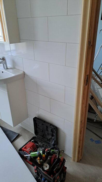 Kaklad vägg i badrum med synlig eldosa utan hål, vit handfat till vänster, verktygsväska i förgrunden.
