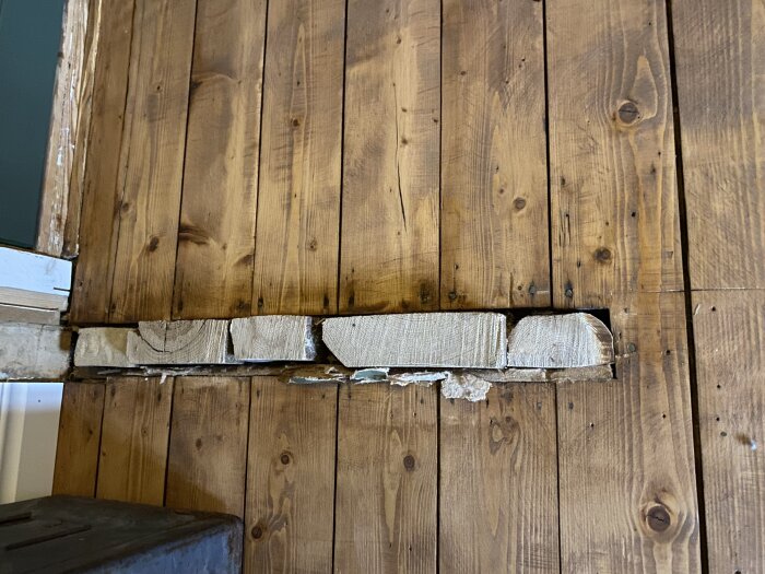 Bild av en delvis demonterad trävägg med synliga träbjälkar och skador där ett skåp har monterats bort.
