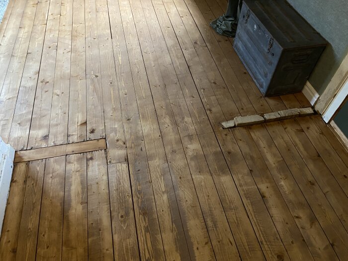 Renoverat trägolv med synlig reparation och gammal trälåda i ett rum, söker timmer för väggåterställning.