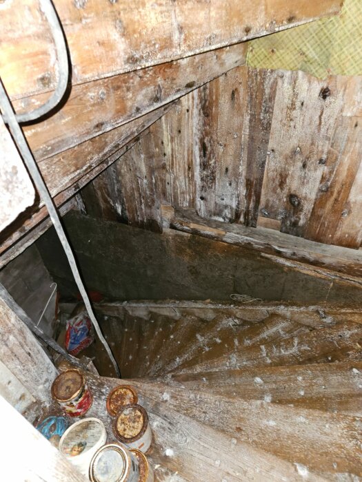 Nedslitna trätrappsteg som leder ned till en fuktig källare med mögel och rostiga burkar på golvet.