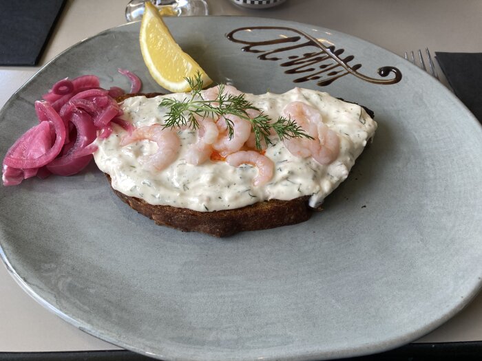 Toast Skagen med räkor, majonnäs och dill, serverad med picklad rödlök och citron på en grå tallrik.