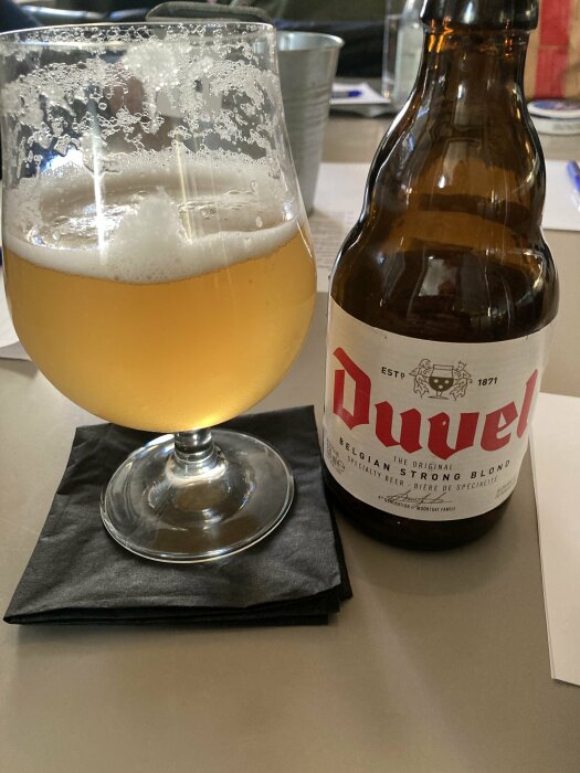En halvtömd ölflaska av märket Duvel bredvid ett glas fyllt med öl på en svart servett.