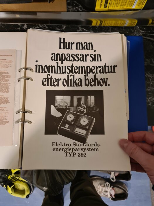 Hand håller en öppen manual med titeln "Hur man anpassar sin inomhustemperatur efter olika behov" över en huspärm.