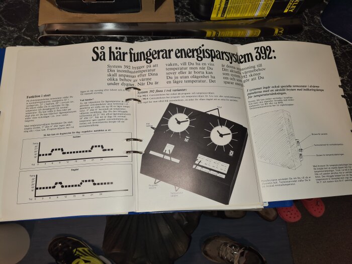 Öppen manual om energisparande system med diagram och illustrationer på ett bord, synliga skor i förgrunden.