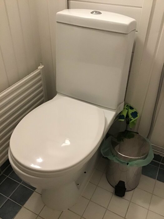 Vit WC-stol i ett badrum med spolknapp på ovansidan, intill en radiator och papperskorg.