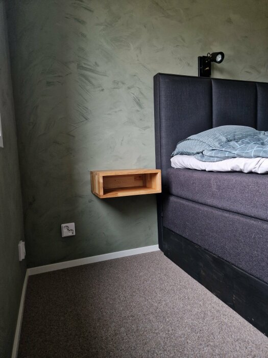 Hörn av ett sovrum med en grå säng och en vägghängd nattduksbord i ek, mot en texturerad grönmålad vägg.