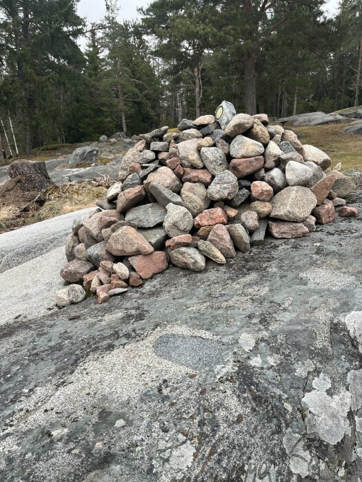 En hög med olika storlekar av runda och kantiga stenar som ligger på fast berggrund i en skogsmiljö.