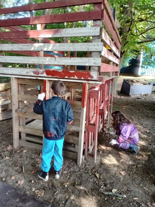 Två barn målar på en träkonstruktion utomhus, pojken står och flickan sitter.