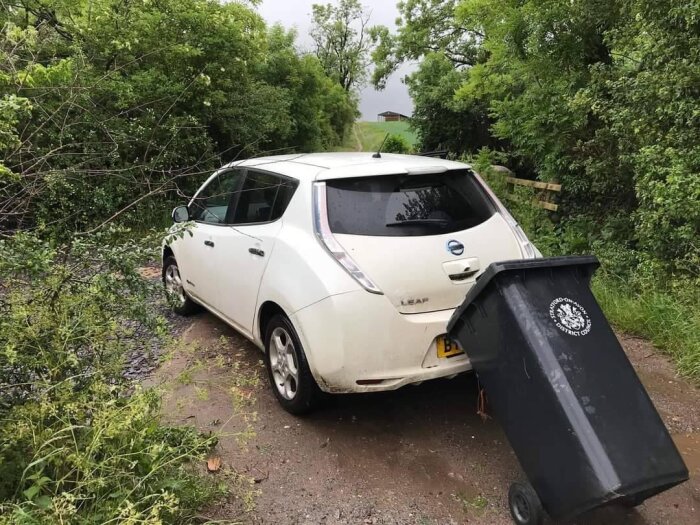 En vit Nissan Leaf parkerad på en grusväg med grenar på taket vid en svart soptunna.