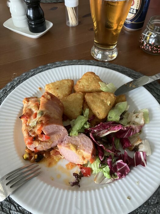 Skivad falukorv i ugn med ost och tomatsås serverad med rösti och blandad sallad på en vit tallrik bredvid ett glas öl.