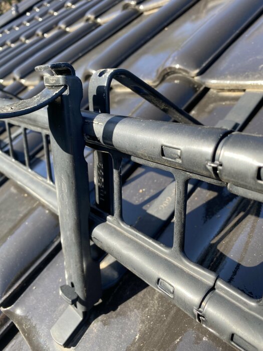 Närbild på en svart snöfångare monterad på takpannor, med synlig låsbygel och delar av monteringsmekanismen.