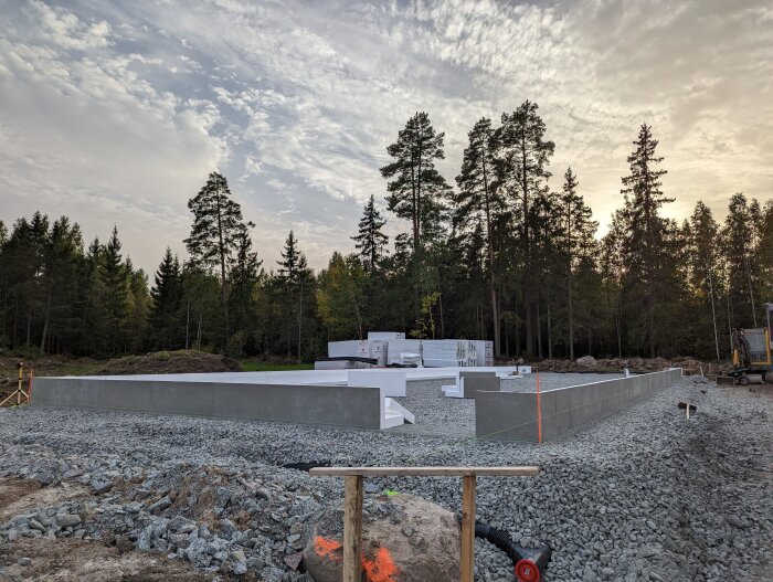 Byggplats med uppsatta kantelement för bostadshus, verktyg och skog i bakgrunden under en molnig himmel.