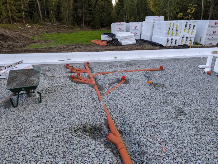 Grundläggning av bostadshus med kantelement och grus, avloppsrör placerade på marken, snören och byggmaterial i bakgrunden.